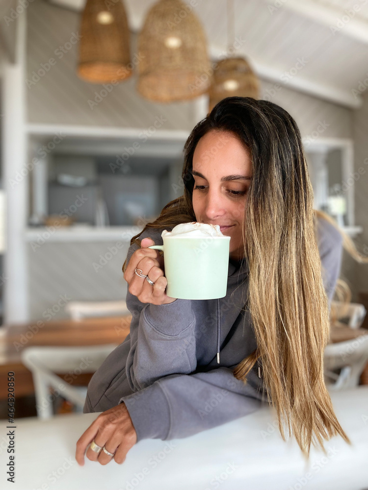 Niña latina tomando una taza de chocolate o café caliente y muy espumoso, lleva poleron gris, pelo aclarado y accesorios como anillos. Su entorno es cálido y claro, se encuentra en el living Niña latina tomando una taza de chocolate o café caliente y muy espumoso, lleva poleron gris, pelo aclarado y accesorios como anillos. Su entorno es cálido y claro, se encuentra en el living