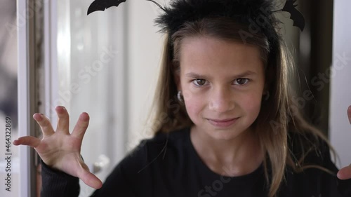 Close-up cute girl scaring looking at camera gesturing appearing from house. Cheerful confident Caucasian child in bat headband posing on Halloween frightening. Slow motion