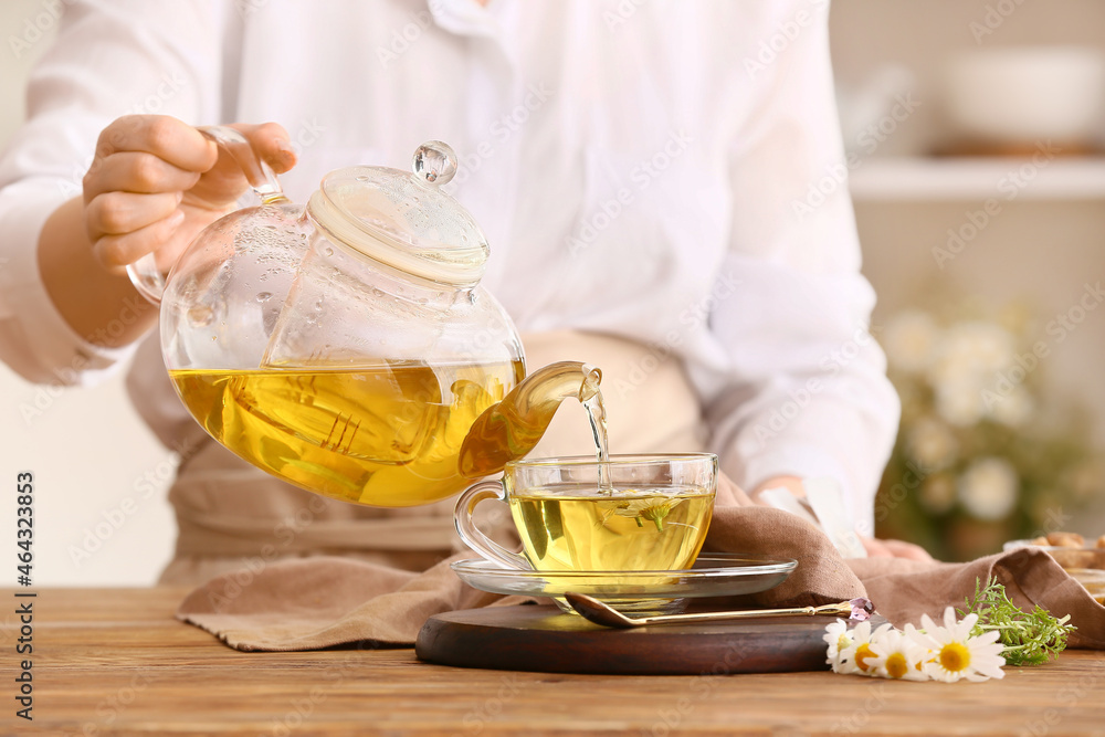 Woman pouring natural chamomile tea from teapot into cup on table Woman pouring natural chamomile tea from teapot into cup on table
