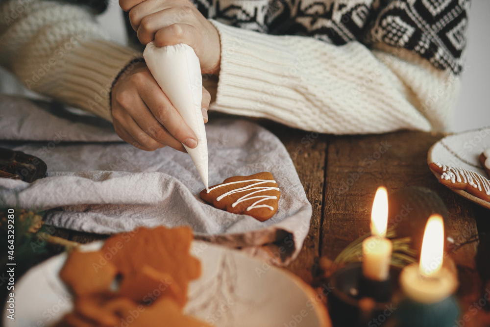 Hands decorating gingerbread cookie christmas tree with frosting on rustic table with napkin, candle, spices, decorations. Atmospheric moody image. Making traditional christmas gingerbread cookies