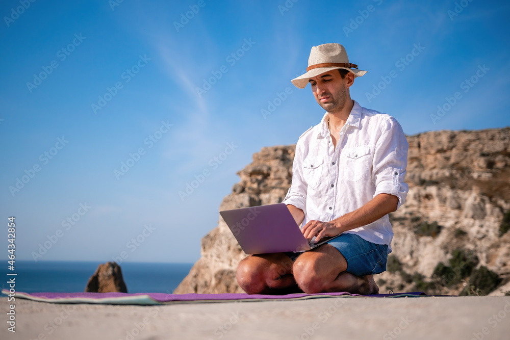 A digital nomad, a man in a hat, a businessman with a laptop does yoga on the rocks by the sea at sunset time, does a business operation online from a distance. Remote work on vacation.