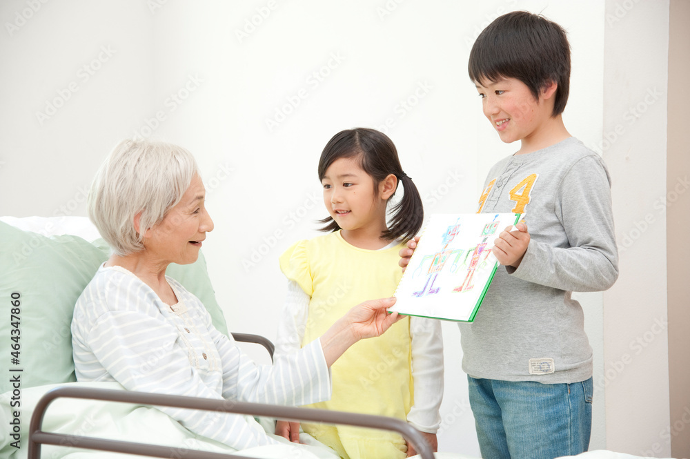 介護ベッドのシニア女性に絵を見せる孫男女