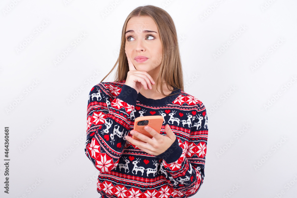 Thinking dreaming Young beautiful caucasian girl wearing christmas sweaters on white background using mobile phone and holding hand on face. Taking decisions and social media concept. Thinking dreaming Young beautiful caucasian girl wearing christmas sweaters on white background using mobile phone and holding hand on face. Taking decisions and social media concept.