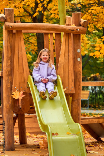 The girl went down the hill. The child plays on the playground in the fall. Wooden platform on the territory of the new house. Happy childhood. Yellow foliage on the trees on a sunny day.
