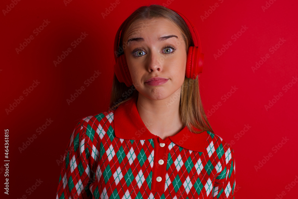 Serious displeased Young beautiful caucasian girl wearing christmas sweaters on red background looks puzzled at camera being angry wears stereo headphones listens music while walking at street Serious displeased Young beautiful caucasian girl wearing christmas sweaters on red background looks puzzled at camera being angry wears stereo headphones listens music while walking at street