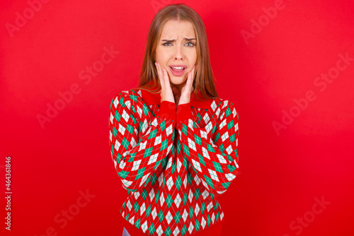 Upset Young beautiful caucasian girl wearing christmas sweaters on red background touching face with two hands © Jihan