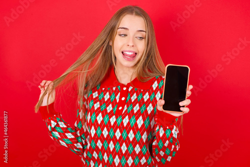 Photo of nice pretty Young beautiful caucasian girl wearing christmas sweaters on red background demonstrate phone screen hold hair tails