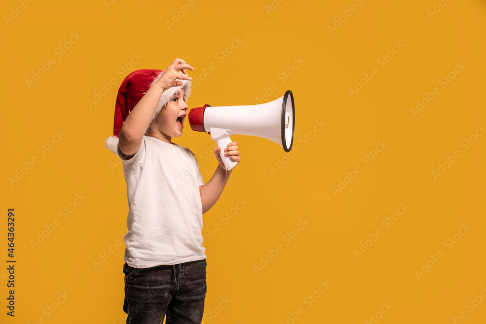 Funny smiling little child boy in Santa hat holding megaphone in hand , smiling and screaming posing on studio background. Funny smiling little child boy in Santa hat holding megaphone in hand , smiling and screaming posing on studio background.