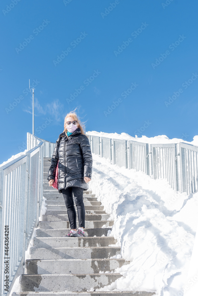 An adult woman wearing a coat, sunglasses and a face mask standing on step of a staircase with a lot of snow around her