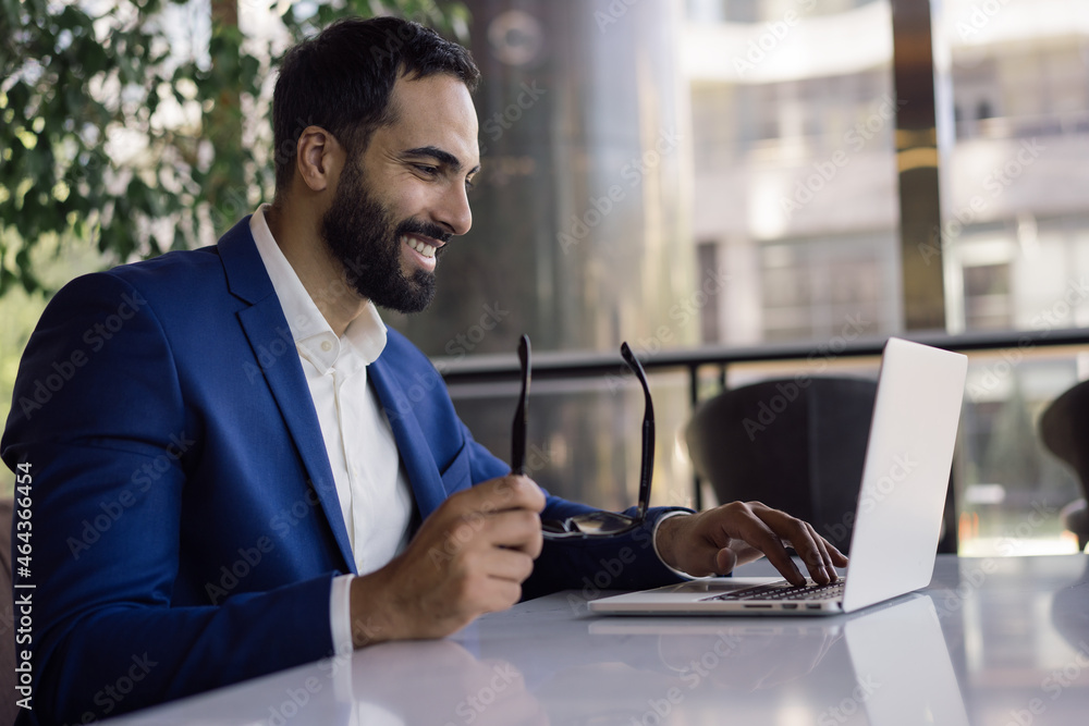 Handsome smiling arabic businessman planning start up project, typing on keyboard in modern office. Confident middle eastern man wearing suit holding eyeglasses working online, sitting at workplace - obrazy, fototapety, plakaty 