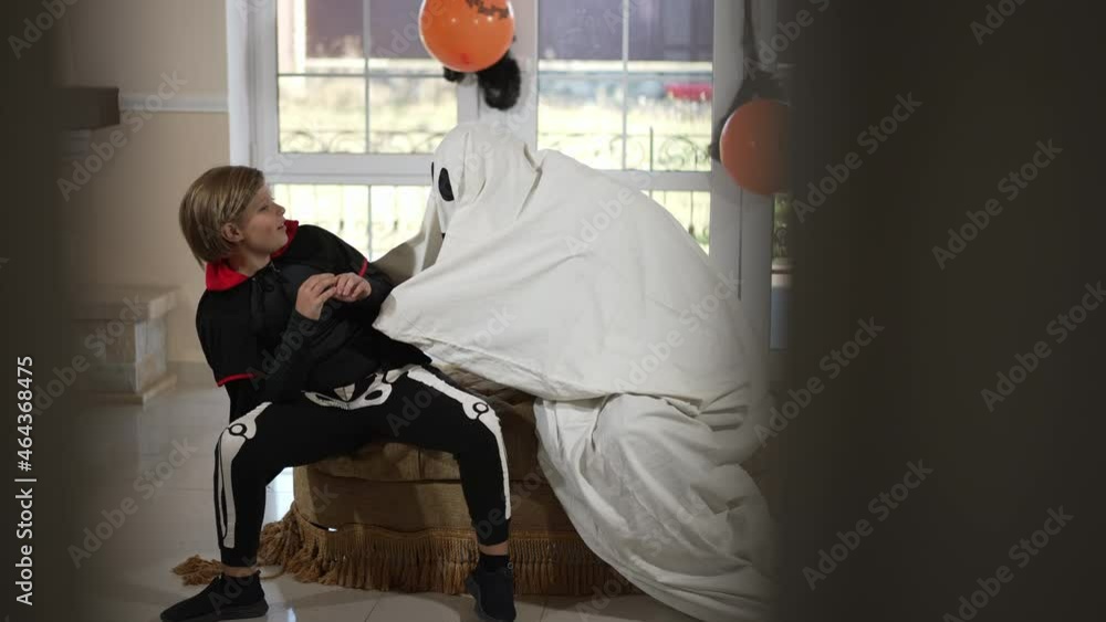 Wide shot scary ghost frightening cute boy sitting in living room on Halloween. Spooky person in costume scaring Caucasian child indoors in slow motion. Traditional celebration and childhood Wide shot scary ghost frightening cute boy sitting in living room on Halloween. Spooky person in costume scaring Caucasian child indoors in slow motion. Traditional celebration and childhood