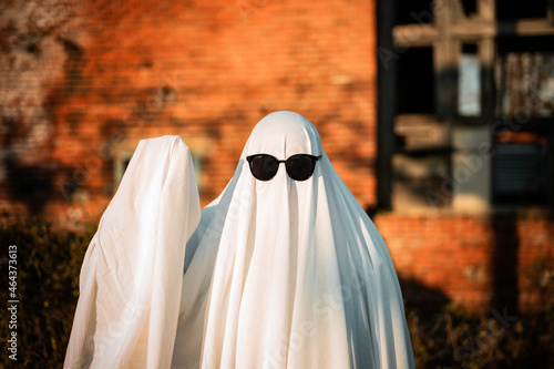 A man in a ghost costume made from a sheet and sunglasses stands near an abandoned building and waves his hand. Ghost Challenge 2021. Spooky season. Celebrating halloween.