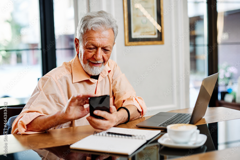 A happy senior student is sitting in a cafe and using his phone to check up on updates related to school. On the table are a laptop and notebook. A happy senior student is sitting in a cafe and using his phone to check up on updates related to school. On the table are a laptop and notebook.