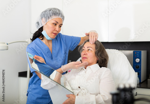 Young female cosmetologist working in the clinic attentive examines the face of an elderly female client before the ..procedure