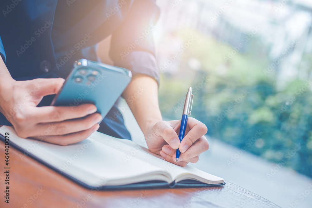 Business woman hand writing on a notepad with a pen and she is holding a cell phone in the office.