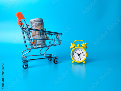 Shopping chart, banknotes and alarm clock on a blue background with copy space.Shopping time and money concept.