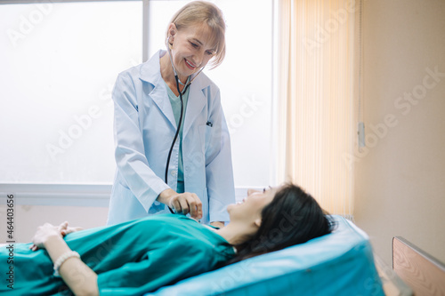 A female doctor examining a patient with a stethoscope while smiling at the patient lying on the bed