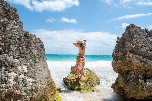 A woman in a flowy orange skirt stands on the rock on the beach in Tulum, white sands and blue water