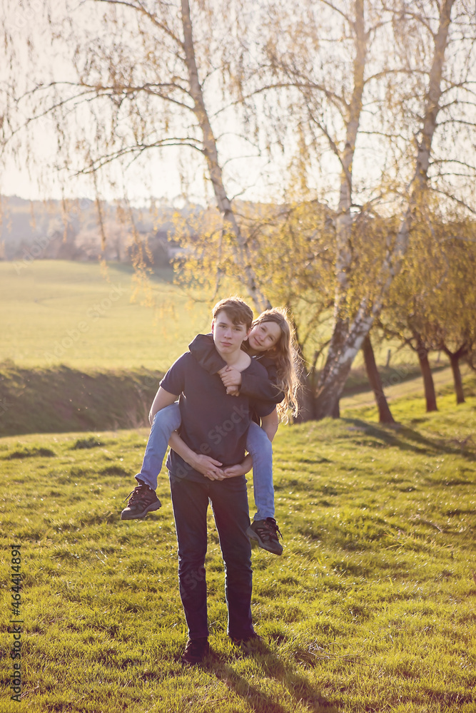 Young boy, preteen, holding his sister on his back. Both are looking at the camera, standing on a field during sunset, with trees behind them, forest far away. Spring, summer, automne. Family moment