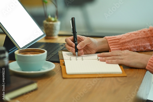 Woman sitting in front of computer tablet and writing information on notebook.