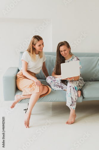 two adorable young women chatting at home on the couch, drinking coffee and working on a laptop