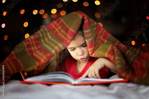 Cute preschool girl reading a book with flashlight on the bed under the covers at night, education and book lovers, lifestyle