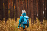 Hiking in pine forest. Woman with sport clothing, backpack and knit hat standing in dry grass and looking at woodland