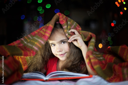 Cute preschool girl reading a book with flashlight on the bed under the covers at night, education and book lovers, lifestyle