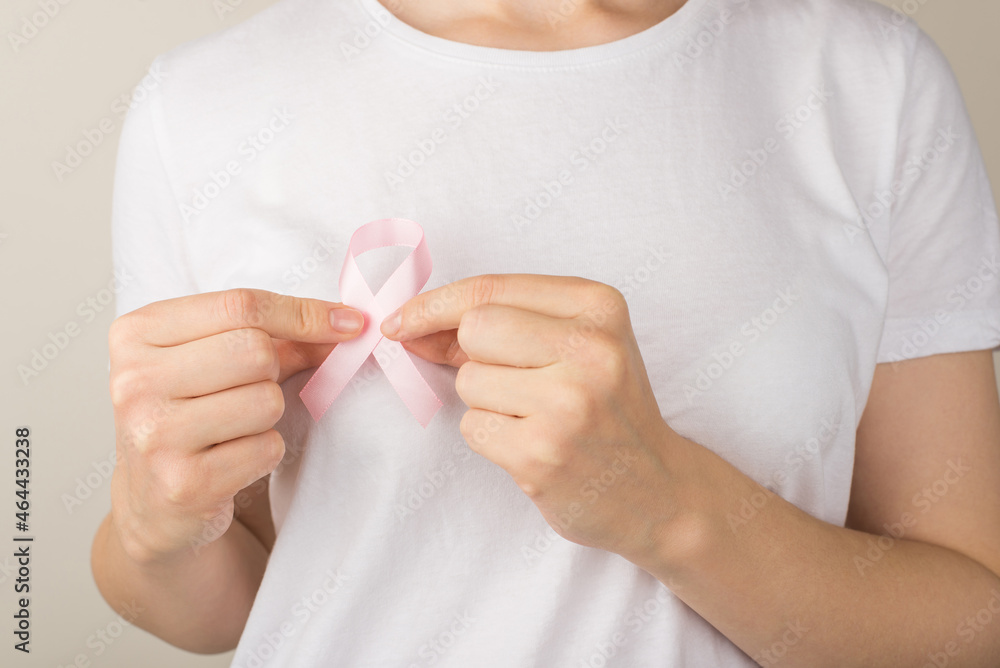 Photo of woman in white t-shirt holding pastel pink ribbon symbol of breast cancer awareness on isolated grey background Photo of woman in white t-shirt holding pastel pink ribbon symbol of breast cancer awareness on isolated grey background