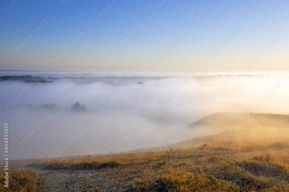 Autumn landscape in the early morning. Fog-covered expanses through which the first rays of the rising sun pass. Trees and hills in the fog. Dawn on a cold autumn morning.