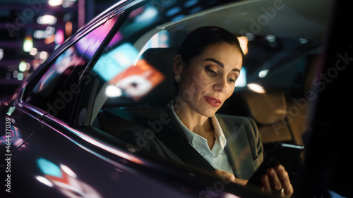 Beautiful Businesswoman is Commuting from Office in a Backseat of Her Car at Night. Entrepreneur Using Smartphone while in Transfer Taxi in Urban City Street with Working Neon Signs. © Gorodenkoff