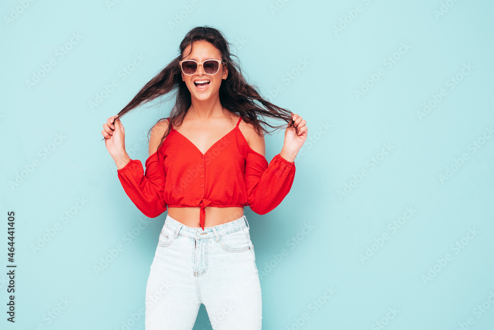 Young beautiful smiling female in trendy summer red top and jeans clothes. Sexy carefree woman posing near blue wall in studio. Positive brunette model having fun. Cheerful and happy. In sunglasses Young beautiful smiling female in trendy summer red top and jeans clothes. Sexy carefree woman posing near blue wall in studio. Positive brunette model having fun. Cheerful and happy. In sunglasses