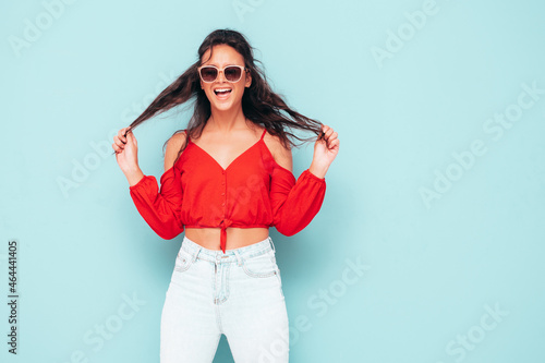 Young beautiful smiling female in trendy summer red top and jeans clothes. Sexy carefree woman posing near blue wall in studio. Positive brunette model having fun. Cheerful and happy. In sunglasses © halayalex