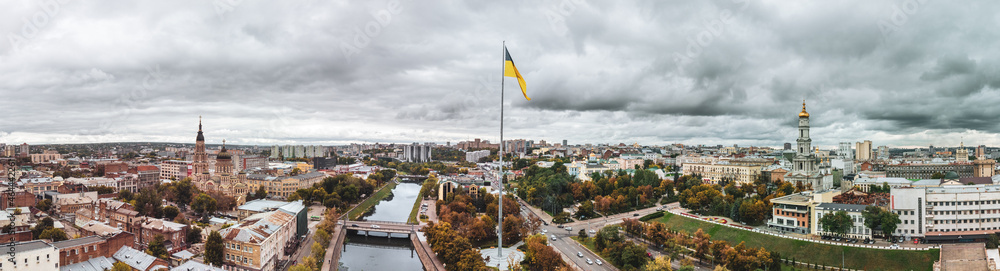 Flag of Ukraine on flagpole with epic heavy cloudscape, wide city aerial panorama on autumn river Lopan embankment, Holy Annunciation Cathedral, Dormition Cathedral in Kharkiv, Ukraine