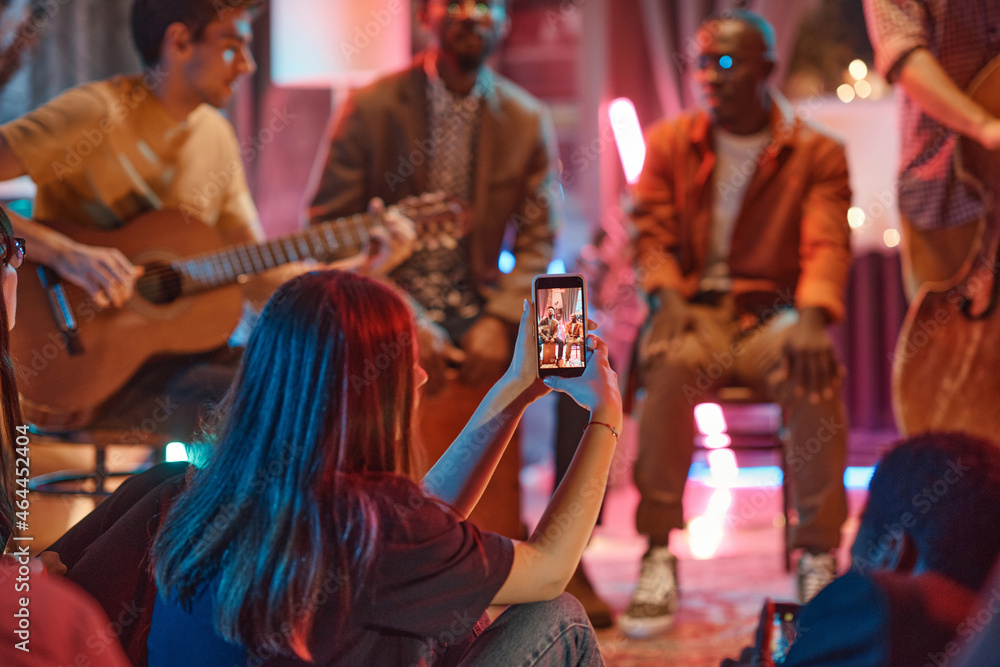 Rear view of young woman shooting the performance of musical band on her mobile phone in the club