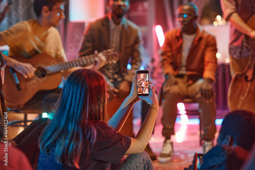 Rear view of young woman shooting the performance of musical band on her mobile phone in the club