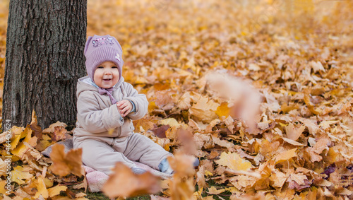 Little cheerful girl sits under a tree in autumn yellow foliage. Copy space