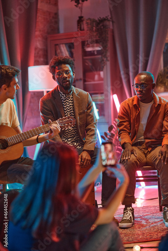 African young man sitting on chair and singing he performing together with his band for people in the club