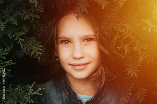 portrait of the face of a cute happy caucasian candid healthy eight year old kid girl surrounded by branches and leaves of green plant thuja or cypress on nature outdoor. flare