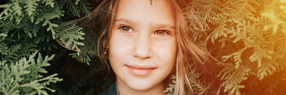 portrait of the face of a cute happy caucasian candid healthy eight year old kid girl surrounded by branches and leaves of green plant thuja or cypress on nature outdoor. banner. flare portrait of the face of a cute happy caucasian candid healthy eight year old kid girl surrounded by branches and leaves of green plant thuja or cypress on nature outdoor. banner. flare