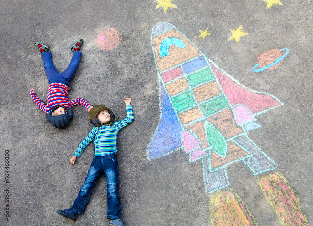 Obraz premium Two little kids boys having fun with drawing universe and space shuttle picture with colorful chalks. Creative leisure for children outdoors in summer. Siblings, friends playing and painting on ground