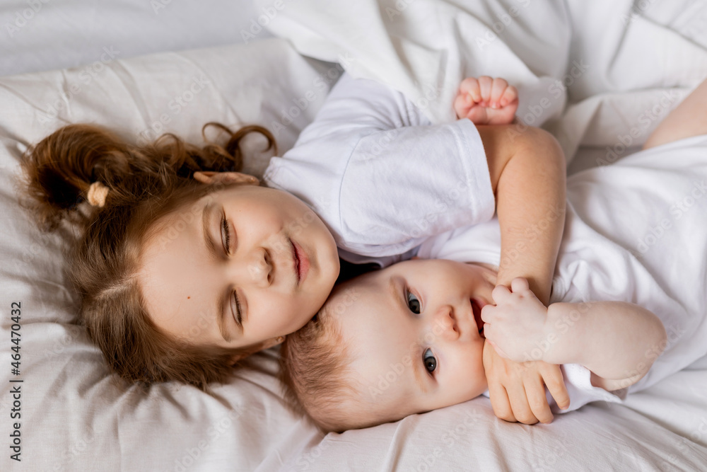 little girl and baby in white clothes are lying in bed on white bed linen. brother and sister bask in bed. children's sleep and rest. hygge, lifestyle. space for text. High quality photo little girl and baby in white clothes are lying in bed on white bed linen. brother and sister bask in bed. children's sleep and rest. hygge, lifestyle. space for text. High quality photo