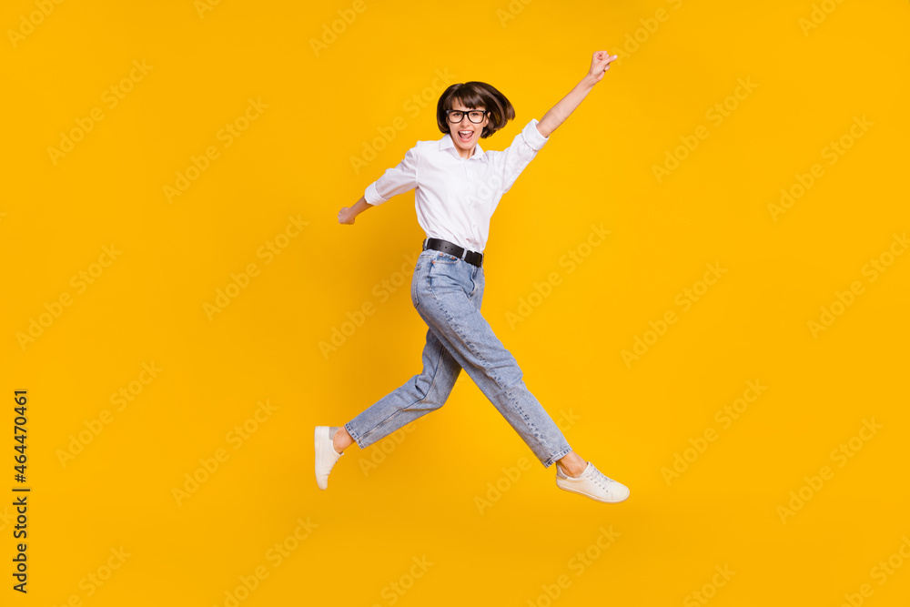 Photo of cheerful hero lady jump raise fist wear specs white shirt jeans sneakers isolated yellow color background