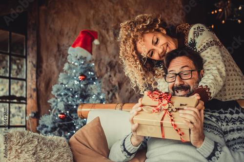 Overjoyed man receive christmas gift from his wife. Happy people enjoy and celebrate christmas eve at home. Xmas tree in background. Winter leisure couple lifestyle indoor