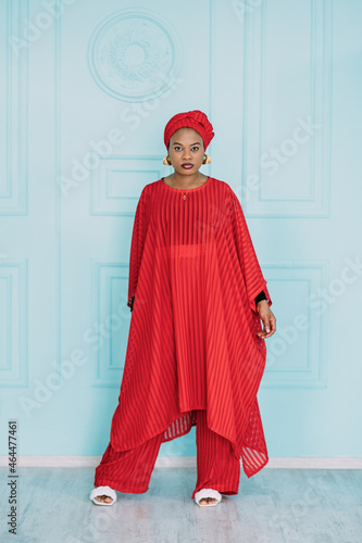 Gorgeous trendy young African American woman in red stylish suit and headscarf, standing against light blue studio wall background