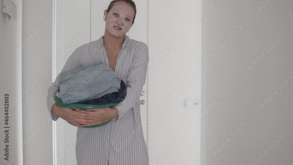 A funny woman with a basin of washed clothes in her hands and a moisturizing mask on her face stands to the hallway. A funny woman with a basin of washed clothes in her hands and a moisturizing mask on her face stands to the hallway.
