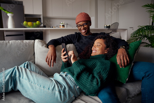 Multi-ethnic couple laughing, looking at text on phone while on the couch in modern apartment