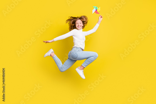 Full length photo of happy nice little girl jump up smile hold hand pinwheel isolated on yellow color background