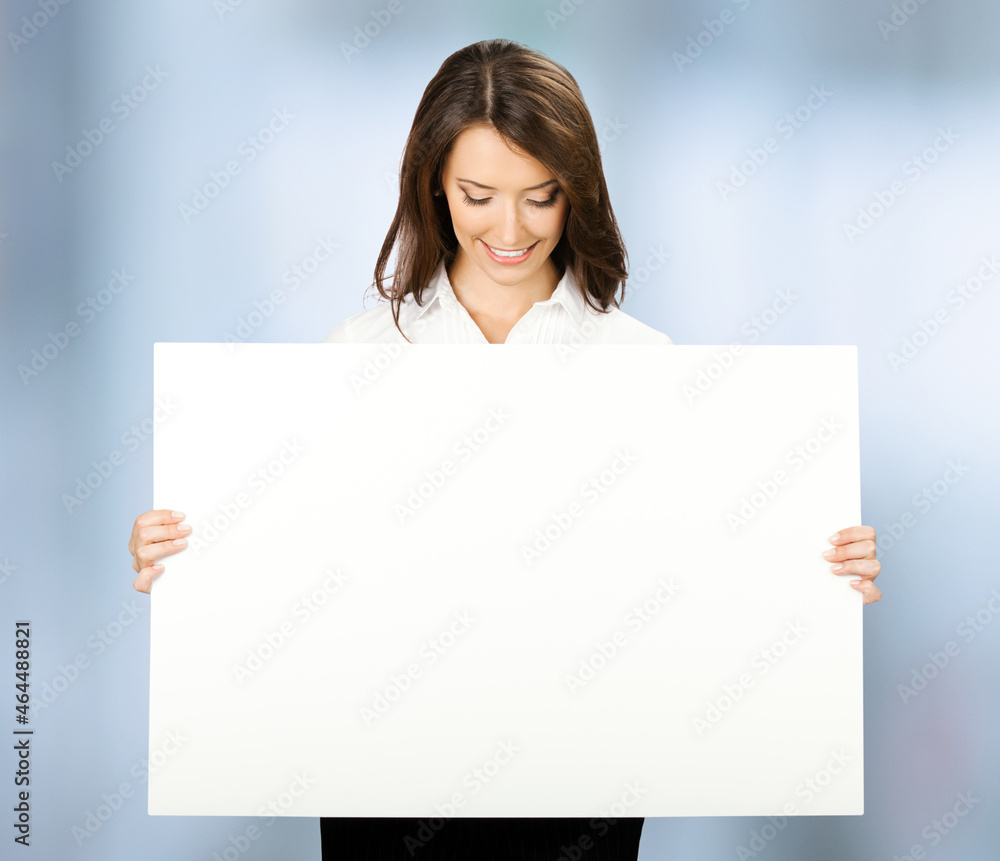 Happy smiling woman holding looking down at mock up empty signboard. Businesswoman in white confident wear - advertising studio concept. Copy space area. Blurred modern office background. Happy smiling woman holding looking down at mock up empty signboard. Businesswoman in white confident wear - advertising studio concept. Copy space area. Blurred modern office background.