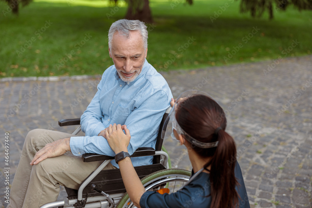 Senior aged man, recovering patient in wheelchair looking and holding hand of nurse wearing face shield and mask, supporting him Senior aged man, recovering patient in wheelchair looking and holding hand of nurse wearing face shield and mask, supporting him